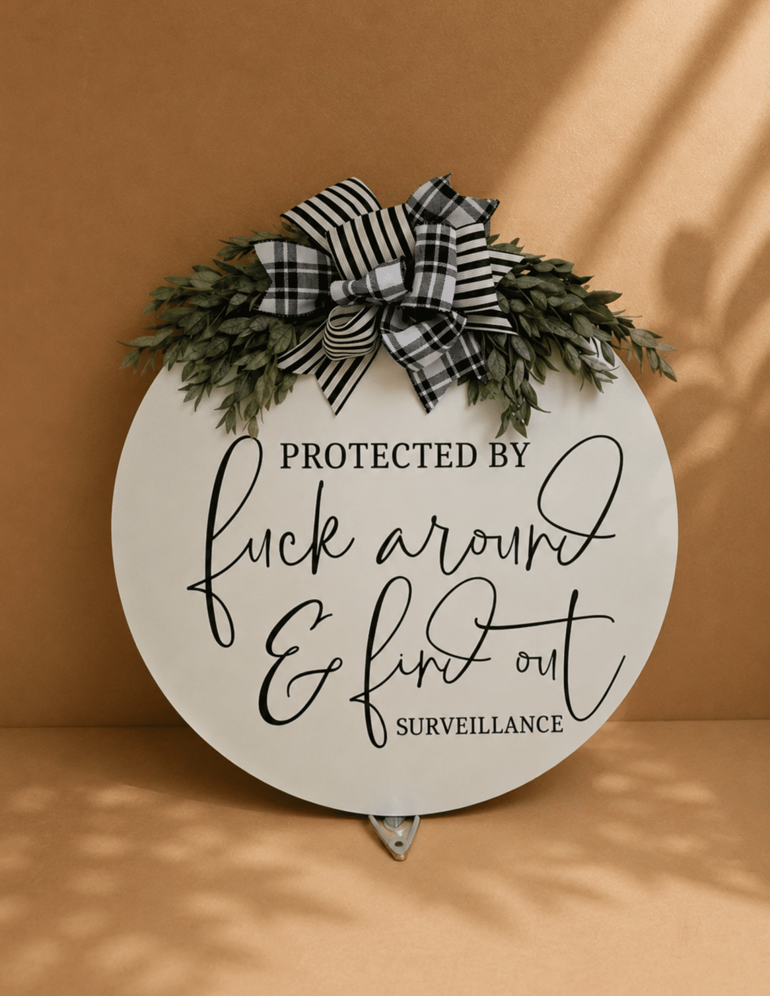 Round Door Sign – Protected by Fuck Around & Find Out Surveillance with modern script, greenery, and decorative plaid bow.