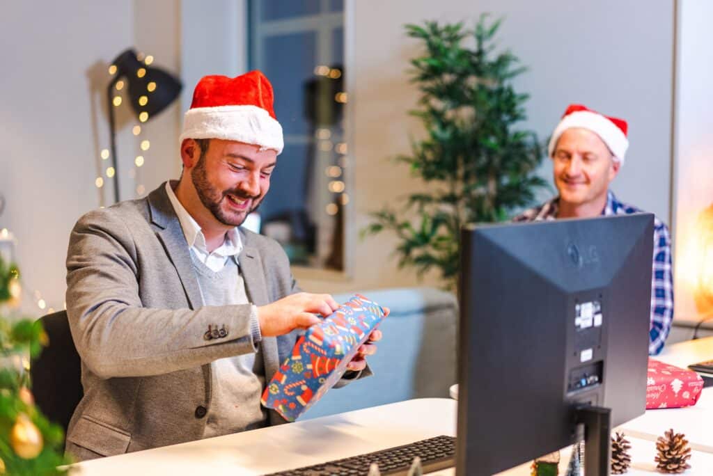Coworker unwrapping a Christmas gift at his office desk while a colleague in a Santa hat smiles in the background