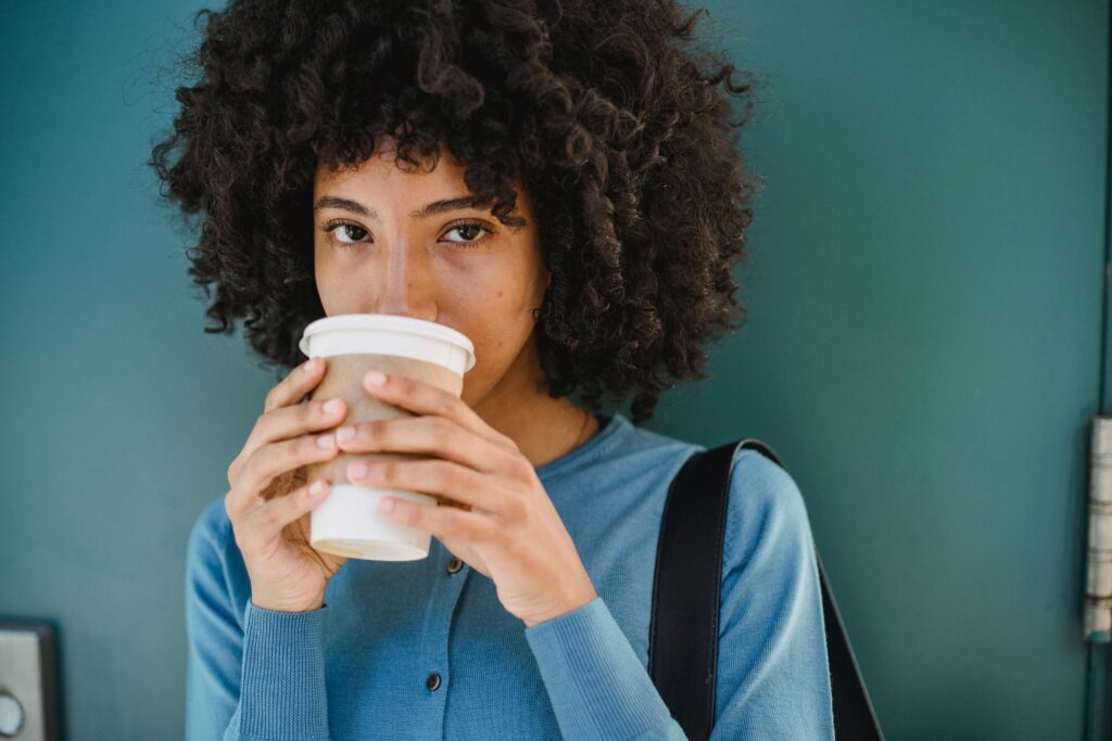 Woman with curly hair drinking coffee from a paper cup against a teal background