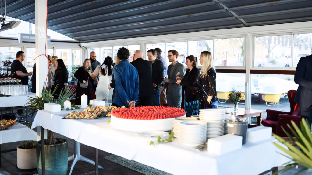 Guests mingling and holding drinks at an indoor corporate event with a catering table of desserts and appetizers